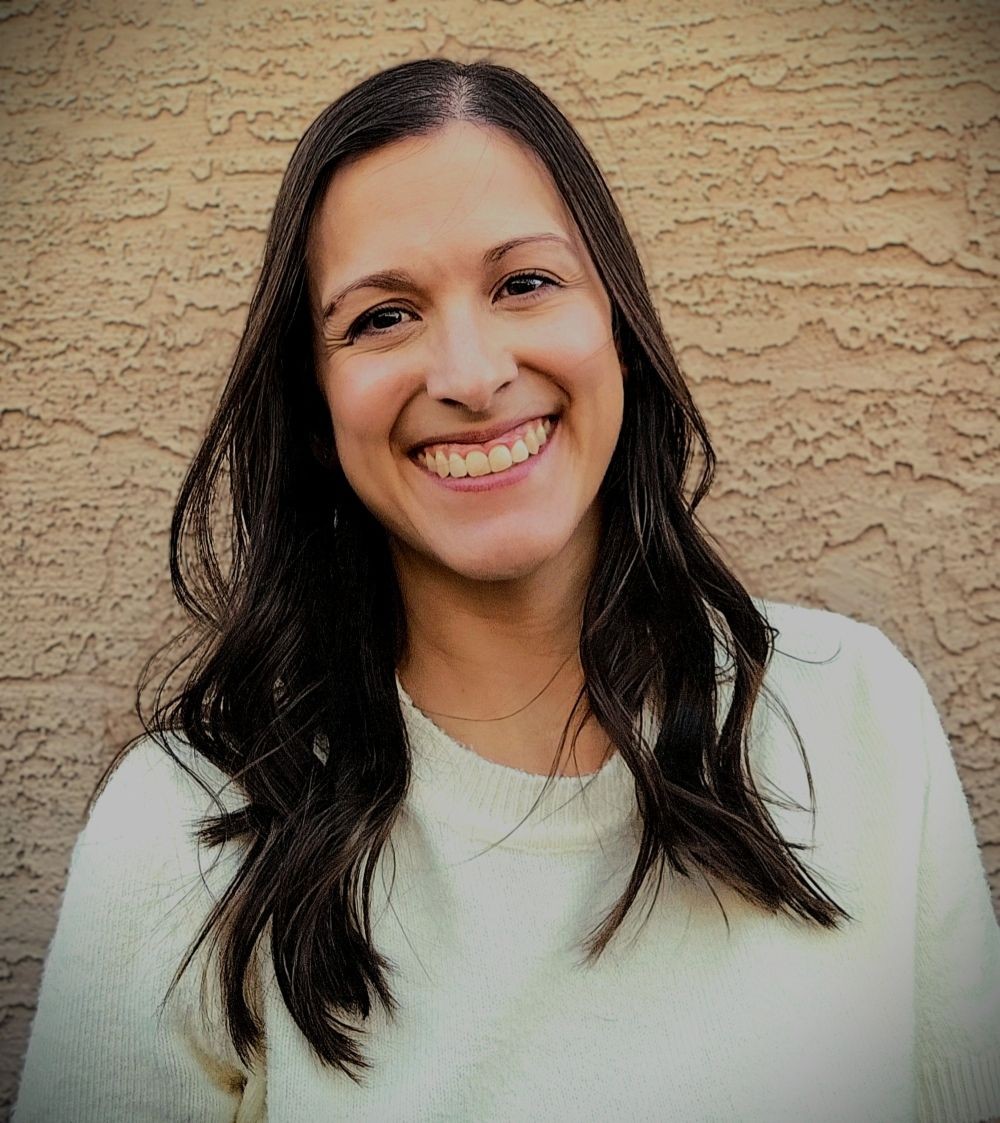 Person wearing a white sweater with dark, wavy hair standing against a textured beige wall.