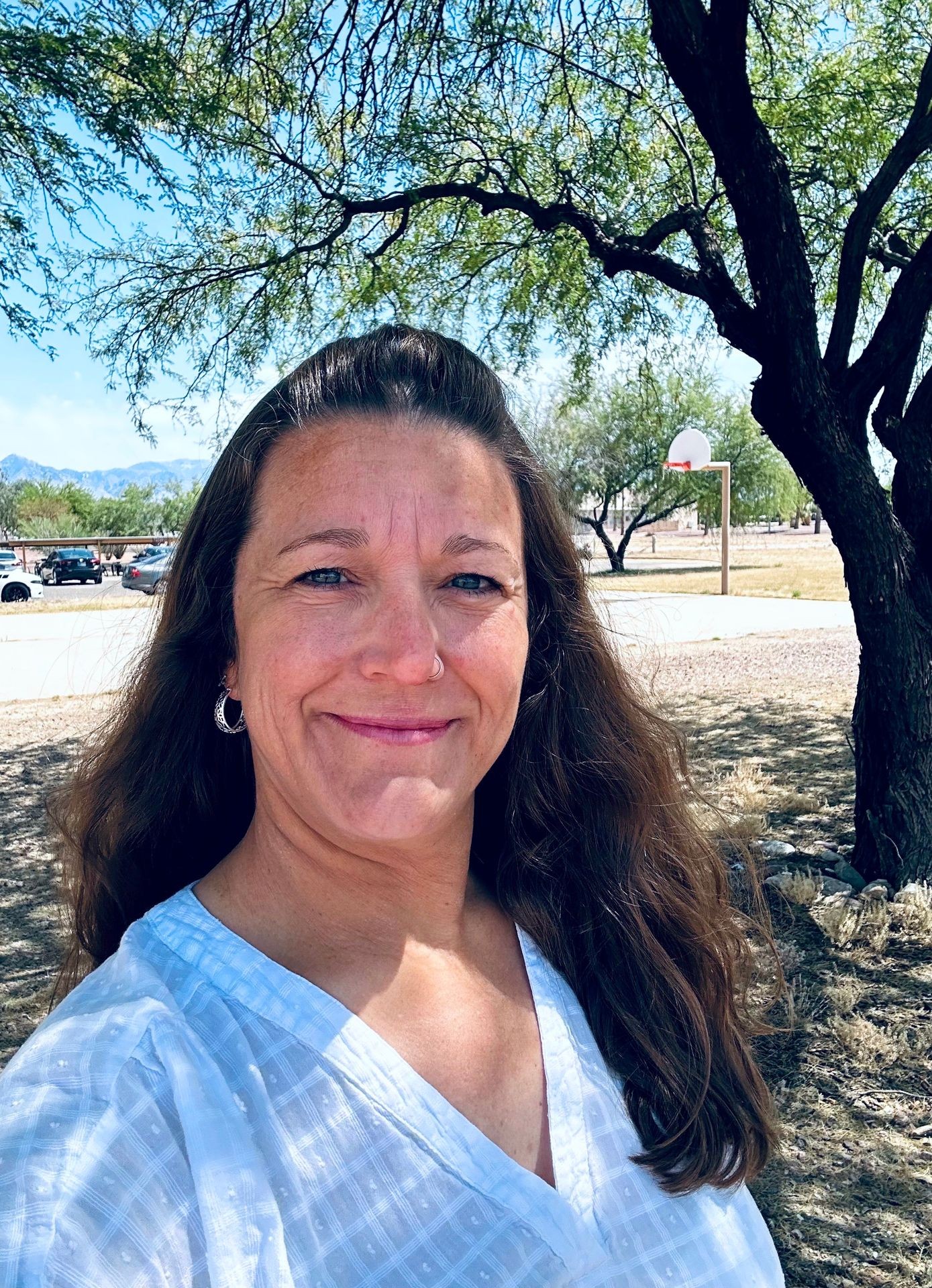 Woman smiling outdoors near a tree and a basketball hoop in a sunny park setting.